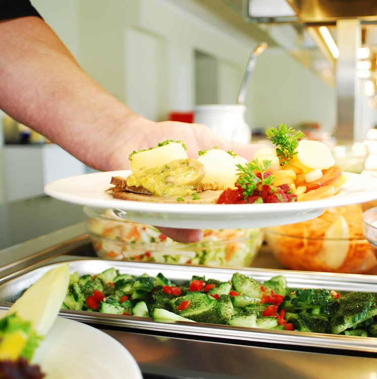 Eine Hand reicht einen frisch angerichteten Teller mit Kartoffeln, Fleisch, Gemüse und Sauce. Im Vordergrund sind Salat- und Gemüsebeilagen an der Essensausgabe sichtbar.