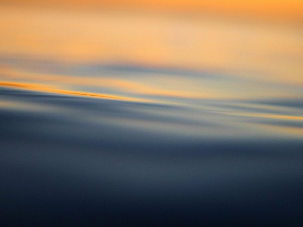 Weiche, fließende Wellen mit sanften Lichtreflexionen in Blau- und Orangetönen, die an einen Sonnenuntergang auf dem Wasser erinnern.